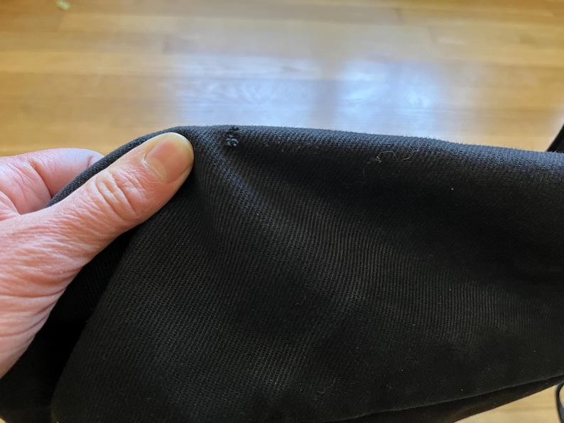 May include: Close-up of black fabric with a small area of damage. The fabric is held by a hand, showing the texture and color of the material. The background is a light brown wooden surface.