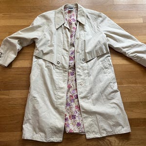 May include: A beige trench coat with a floral patterned lining. The coat has a classic design with a collar, button closures, and a belt. The sleeves have button details at the cuffs. The coat is laid flat on a wooden surface.