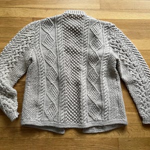 May include: A light gray, hand-knitted cardigan with intricate cable and textured stitch patterns. The sweater features a high collar and long sleeves. The garment is displayed on a wooden surface.