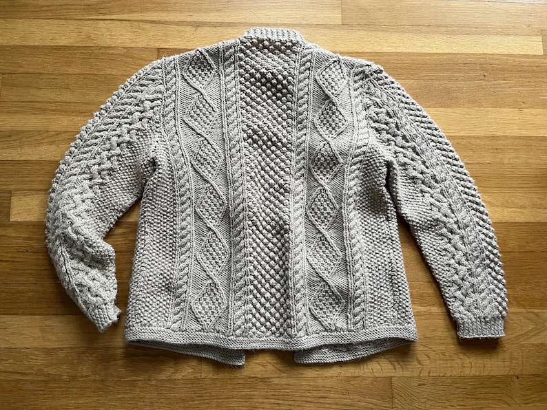 May include: A light gray, hand-knitted cardigan with intricate cable and textured stitch patterns. The sweater features a high collar and long sleeves. The garment is displayed on a wooden surface.