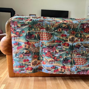 May include: A colorful fabric throw draped over a brown leather sofa. The throw features a detailed illustration of a rural autumn scene with red barns, pumpkins, and apple orchards. The color palette includes shades of red, green, and brown.