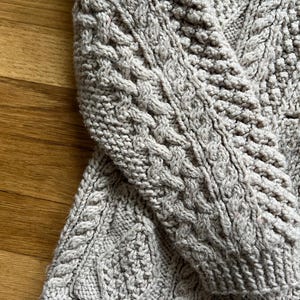 May include: Close-up of a light gray cable-knit sweater. The sweater features intricate cable patterns and a textured knit design. The sleeve is visible, showcasing the detailed craftsmanship of the garment. The background is a wooden surface.