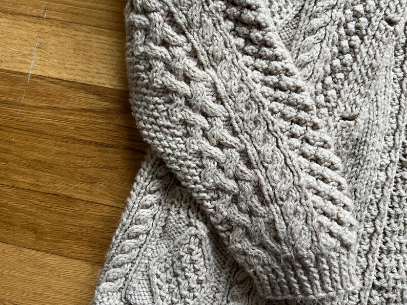 May include: Close-up of a light gray cable-knit sweater. The sweater features intricate cable patterns and a textured knit design. The sleeve is visible, showcasing the detailed craftsmanship of the garment. The background is a wooden surface.