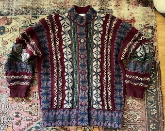 May include: A button-up cardigan sweater in a mix of burgundy, navy, green, and cream colors. The sweater features a high collar and a patterned design with vertical stripes and geometric shapes. The sleeves and cuffs are a solid burgundy color.