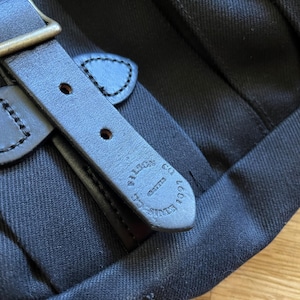 May include: Close-up of a black canvas bag with a leather strap and buckle. The leather strap is dark brown with visible stitching and the words "FILSON CO. EST. 1897" embossed on it. The buckle is brass-colored.