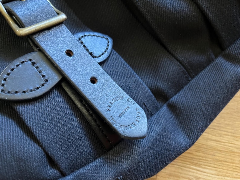 May include: Close-up of a black canvas bag with a leather strap and buckle. The leather strap is dark brown with visible stitching and the words "FILSON CO. EST. 1897" embossed on it. The buckle is brass-colored.