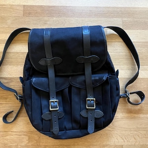 May include: A black backpack with leather straps and brass buckles. The backpack has two front pockets and adjustable shoulder straps. The bag is made of a durable canvas material and is designed for carrying items.