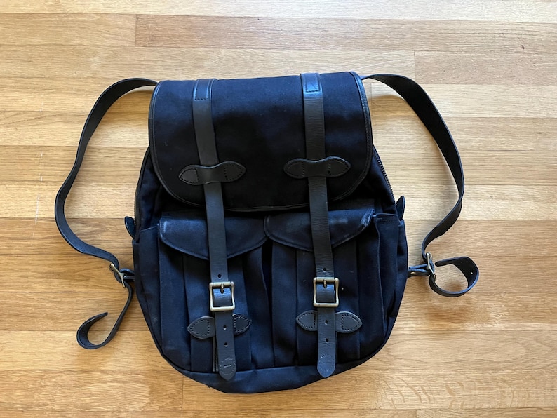 May include: A black backpack with leather straps and brass buckles. The backpack has two front pockets and adjustable shoulder straps. The bag is made of a durable canvas material and is designed for carrying items.