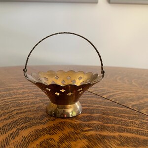 May include: A small, vintage brass basket with a scalloped rim and a handle. The basket has a decorative, openwork design and sits on a round base. The handle is made of twisted wire. The basket is likely used for decorative purposes.