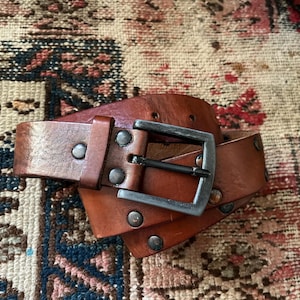 May include: A brown leather belt with a silver-colored buckle and rivets. The belt is made of a thick, textured leather and has several holes for adjustment. The buckle is square and has a simple design. The belt is lying on a patterned rug.