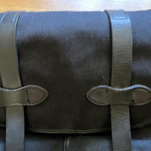 May include: Close-up of a black canvas bag with leather straps and closures. The bag has a rectangular shape with a curved top flap. The leather straps are secured with decorative, stitched leather accents.