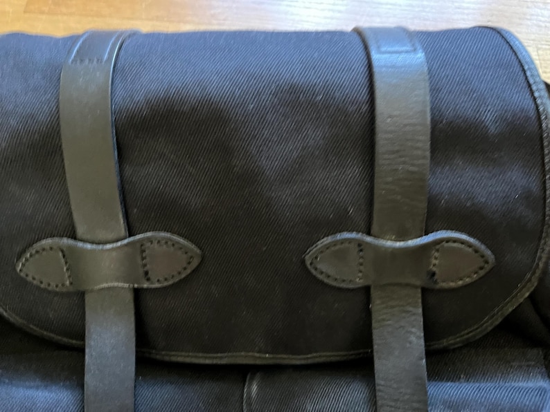 May include: Close-up of a black canvas bag with leather straps and closures. The bag has a rectangular shape with a curved top flap. The leather straps are secured with decorative, stitched leather accents.