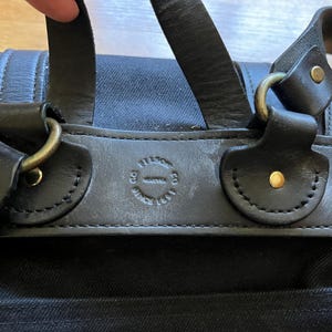 May include: Close-up of a black canvas bag with leather straps and brass hardware. The bag features a leather patch with embossed text. The straps are attached with brass rings and secured with brass rivets.