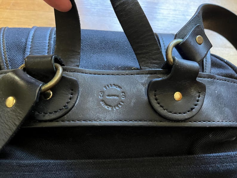 May include: Close-up of a black canvas bag with leather straps and brass hardware. The bag features a leather patch with embossed text. The straps are attached with brass rings and secured with brass rivets.