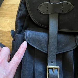 May include: Black canvas messenger bag with a leather strap and buckle closure. The bag features a flap with a leather accent and a shoulder strap. The bag has a pocket on the front and is made of durable material.