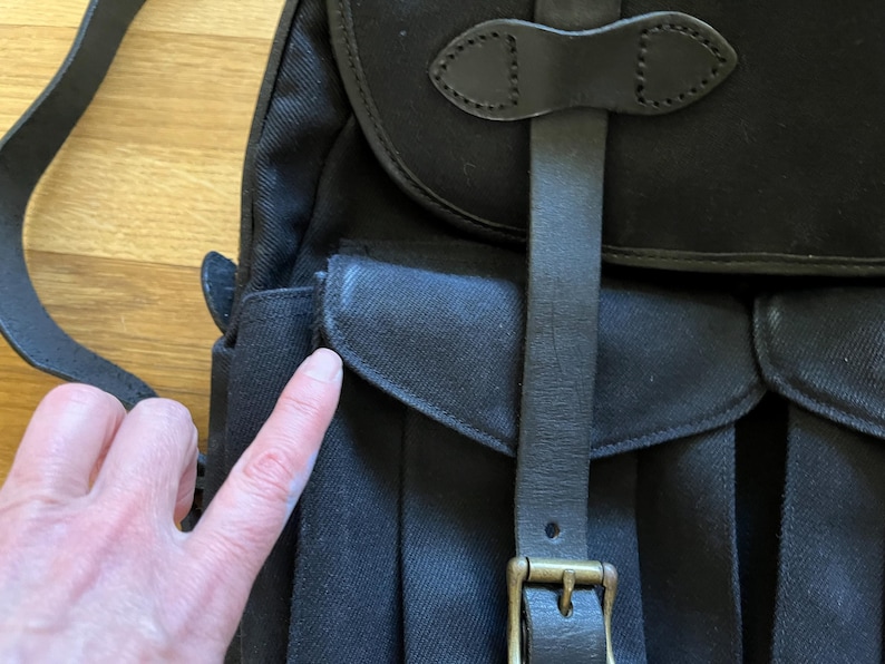 May include: Black canvas messenger bag with a leather strap and buckle closure. The bag features a flap with a leather accent and a shoulder strap. The bag has a pocket on the front and is made of durable material.