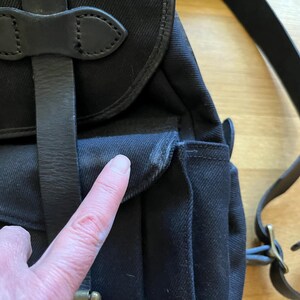 May include: Close-up of a black canvas backpack with leather straps and accents. The bag features a flap closure and adjustable shoulder straps. The leather straps are secured with metal buckles.