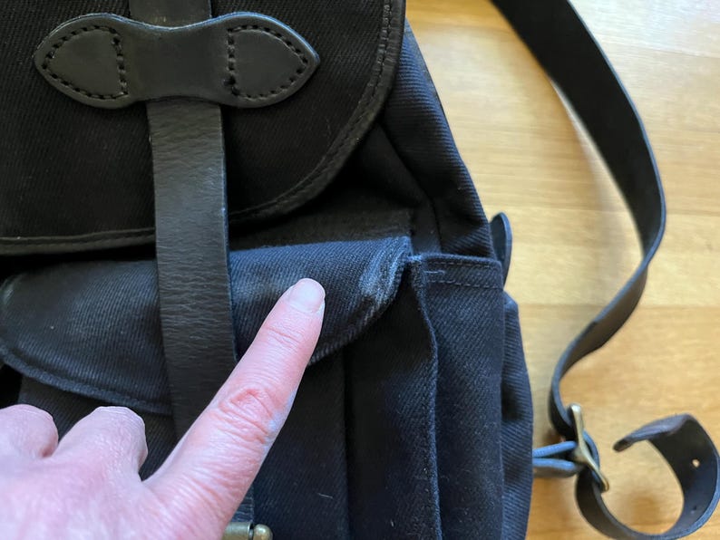 May include: Close-up of a black canvas backpack with leather straps and accents. The bag features a flap closure and adjustable shoulder straps. The leather straps are secured with metal buckles.