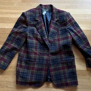 May include: A plaid blazer in shades of burgundy, navy, and olive green. The jacket has a single-button closure, notched lapels, and two front pockets. The blazer is made of a woven fabric and is laid flat on a wooden surface.