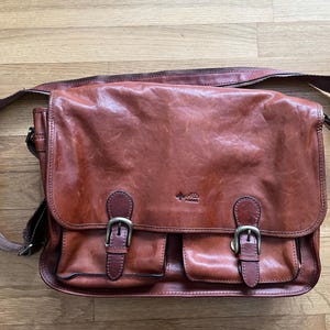 May include: A brown leather messenger bag with a flap closure and two front pockets. The bag has a shoulder strap and metal buckles. The leather has a distressed appearance.