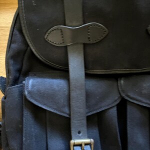 May include: Black canvas backpack with leather straps and buckle closures. The backpack features a flap top and two front pockets with rounded flaps. The leather straps and buckles are a dark brown color.