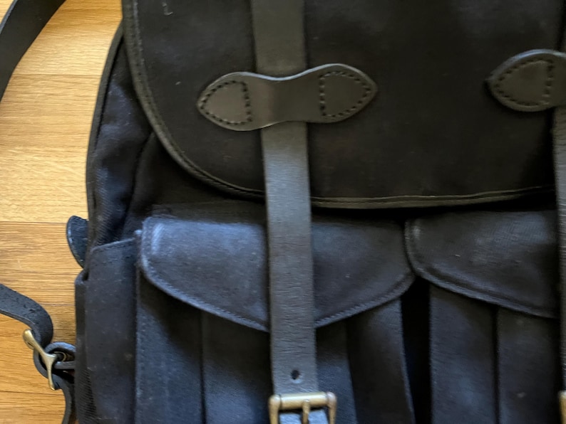 May include: Black canvas backpack with leather straps and buckle closures. The backpack features a flap top and two front pockets with rounded flaps. The leather straps and buckles are a dark brown color.