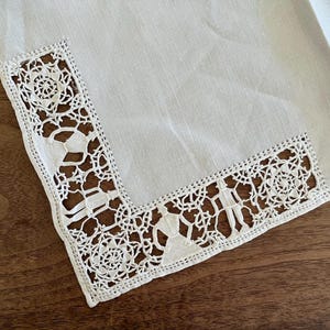 May include: A white linen napkin with a decorative, openwork lace border. The lace features floral and geometric patterns. The napkin is folded, showing a corner with the detailed lace design. The background is a wooden surface.