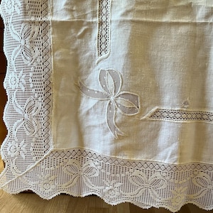 May include: Cream-colored tablecloth with intricate lace detailing. The edges feature a scalloped design with floral and bow motifs. A large bow is embroidered on the tablecloth. The tablecloth is made of a light, possibly linen, fabric.