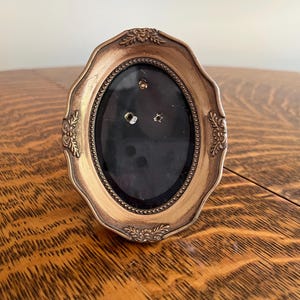 May include: An antique gold-toned oval picture frame with a black interior. The frame has a decorative, scalloped edge and floral accents at the corners. The frame is empty and ready for a photo.