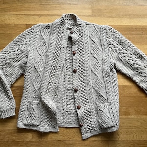 May include: A light gray, cable-knit cardigan with a button-up front and two front pockets. The sweater features intricate knit patterns and brown buttons. The long sleeves and collar are also visible.