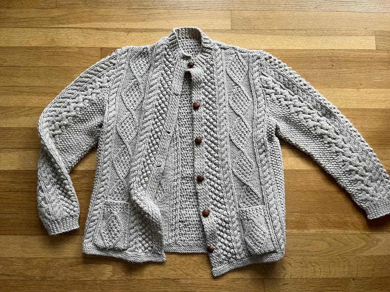 May include: A light gray, cable-knit cardigan with a button-up front and two front pockets. The sweater features intricate knit patterns and brown buttons. The long sleeves and collar are also visible.