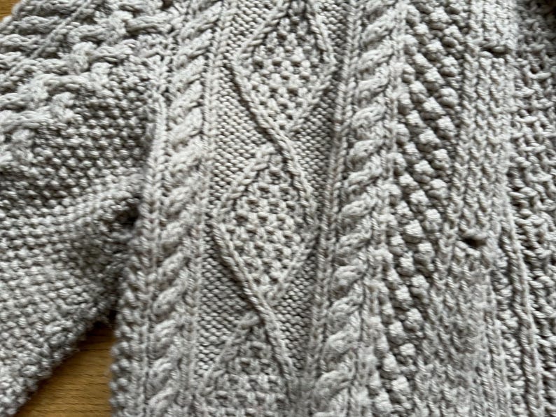 May include: Close-up of a light gray knitted sweater. The sweater features a detailed cable knit pattern with diamond and textured stitch designs. The yarn appears soft and the overall texture is chunky.