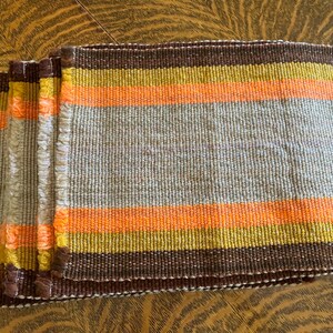 May include: A stack of woven rectangular placemats. Each placemat features horizontal stripes in shades of brown, yellow, orange, and gray. The edges have a fringed texture, adding a rustic touch. These placemats are designed for dining and home decor.