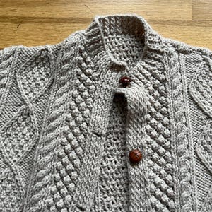 May include: A light gray, hand-knitted cardigan with a textured cable-knit design. The sweater features a collared neckline and is fastened with three brown, round buttons. The intricate knitwork and neutral color make it a versatile garment.