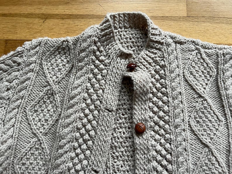 May include: A light gray, hand-knitted cardigan with a textured cable-knit design. The sweater features a collared neckline and is fastened with three brown, round buttons. The intricate knitwork and neutral color make it a versatile garment.