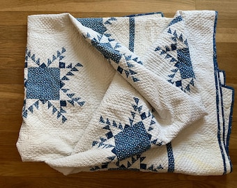 Könnte beinhalten: Eine gefaltete weiß-blaue Steppdecke mit einem sich wiederholenden geometrischen Muster. Die Steppdecke hat ein quadratisches Design mit blauen Akzenten und einer strukturierten Oberfläche. Die Steppdecke ist auf einer Holzoberfläche gefaltet.