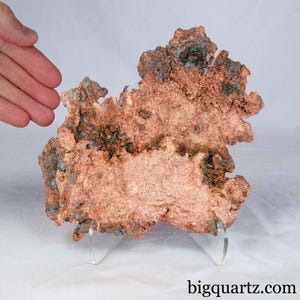 May include: A raw, metallic copper specimen with a jagged, irregular shape and a textured, reddish-brown surface. The mineral features dark, oxidised patches and sits on a clear acrylic display stand. The text bigquartz.com is visible.