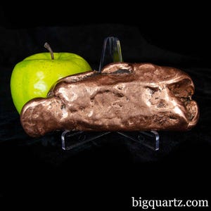 Könnte beinhalten: Ein großes, unregelmäßig geformtes Kupferstück, präsentiert auf einem transparenten Ständer, neben einem Granny Smith Apfel. Das Kupfer hat eine satte, rotbraune Farbe und eine strukturierte Oberfläche. Der Apfel ist hellgrün. Der Hintergrund ist schwarz.