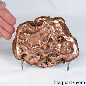 May include: A large, irregular copper nugget with a polished, metallic reddish-brown surface and deep, textured crevices. The specimen is displayed on a clear acrylic stand. The text bigquartz.com is printed in the bottom right corner.