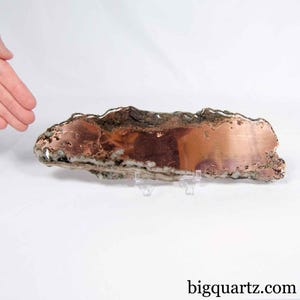 Peut inclure: Une plaque polie de spécimen minéral de cuivre natif présentant une surface métallique brun rougeâtre avec des inclusions naturelles de quartz blanc et gris le long des bords irréguliers, posée sur un support en plastique transparent.