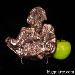 May include: A large, irregularly shaped copper specimen with a textured surface, displayed on a clear stand. A green apple is positioned to the right of the copper. The background is black. The website address "bigquartz.com" is visible at the bottom.