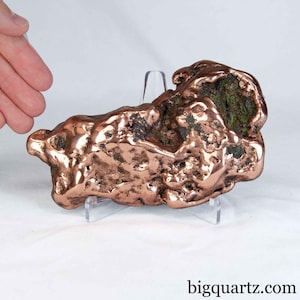 May include: A large, raw copper nugget with a metallic, reddish-brown luster and irregular, pitted surface texture. The specimen features deep crevices and dark, oxidized mineral inclusions, displayed on a clear acrylic stand.