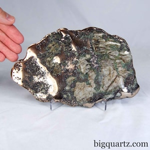 May include: A large, irregular mineral specimen featuring a polished metallic bronze-colored edge surrounding a textured surface of white, green, and dark gray crystalline rock. The piece is displayed on a clear stand with the text bigquartz.com.
