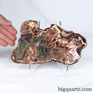 May include: A raw copper specimen featuring a metallic, reddish-brown surface with irregular, textured formations and patches of dark green oxidation. The mineral piece is displayed on a clear acrylic stand, with the text bigquartz.com visible.
