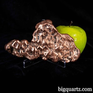 May include: A copper nugget with a textured surface, displayed on a clear stand, next to a green apple. The nugget has a rough, irregular shape and a reddish-brown color. The website "bigquartz.com" is visible in the bottom right corner.