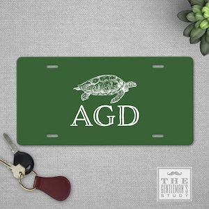 May include: Green license plate with a white turtle illustration and the letters "AGD". The plate has pre-drilled holes for mounting. A set of keys with a leather fob is in the foreground.