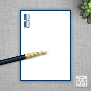 May include: A white notepad with a dark blue border and the letters "A H B" in dark blue in the top left corner. A gold and black pen is lying on the notepad. The text "THE GENTLEMAN'S STUDY" is in a small rectangle in the bottom right corner.
