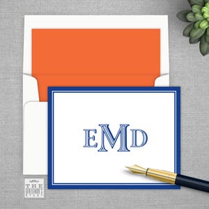 May include: A stationery set featuring a white card with a blue border and monogrammed initials, inside a white envelope with an orange interior. A gold-tipped pen rests nearby. The Gentleman's Study logo is visible.
