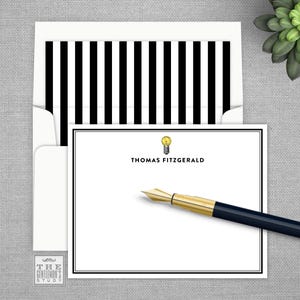 May include: A white card with black stripes on the envelope. The card has a black border and a light bulb graphic with the text "THOMAS FITZGERALD". A gold and black pen is lying on the card.
