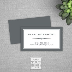 May include: A gray business card with white trim and text. The text reads "Henry Rutherford", a phone number, and an email address. The card also has a small decorative design above the phone number. Below the card is a logo that reads "The Gentleman's Study" with a stylized mustache.